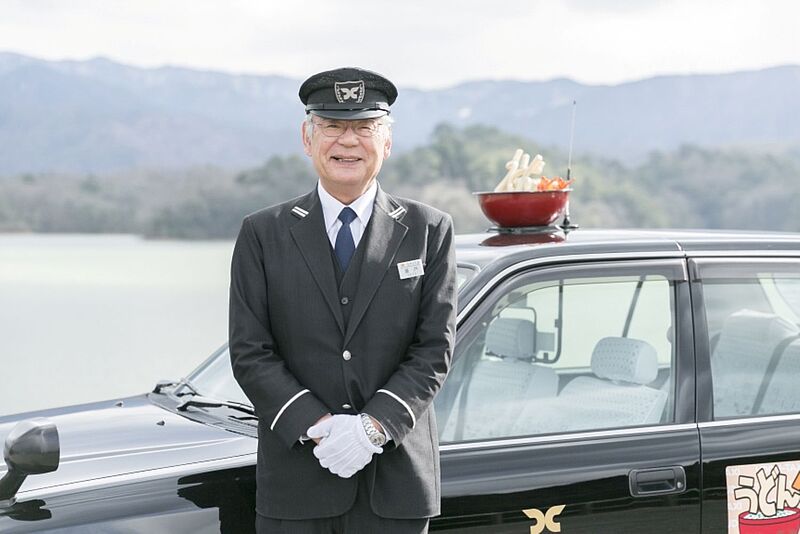 Lächelnder Chauffeur in schwarzer Uniform mit Mütze steht vor dunkler Limousine in Berglandschaft