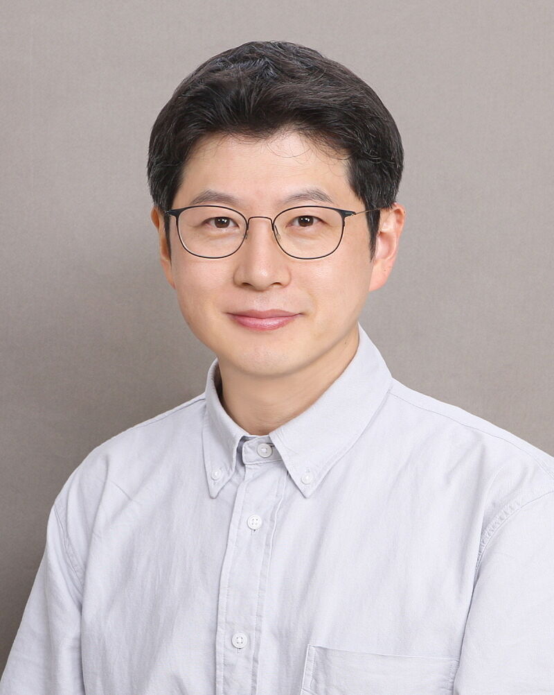 Porträt eines lächelnden asiatischen Mannes mit Brille und weißem Hemd vor grauem Hintergrund.