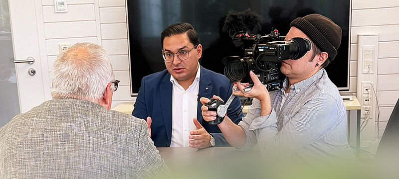 Ein Mann mit schwarzem Jacket und weißem Hemd sitzt mit zwei weiteren Männern an einem Tisch. Ein Mann mit grauen Haaren sitzt mit dem Rücken zum Fotografen, ein anderer hält eine Kamera in der Hand und filmt die Situation. 