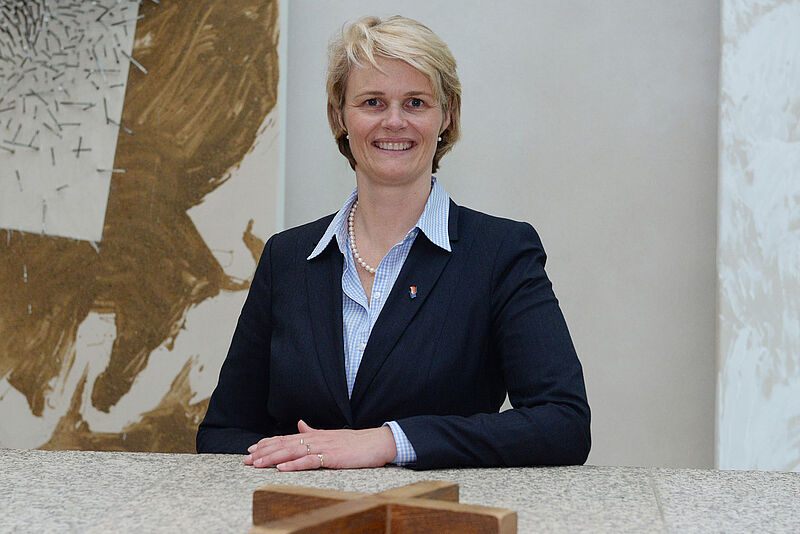 Porträtfoto einer lachenden Frau mit kurzen, blonden Haaren, einer hellblauen Bluse und einem dunkelblauen Blazer. Vor ihr auf dem Schreibtisch liegt ein großes Kreuz aus Holz.