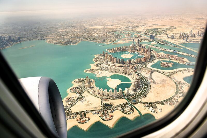 Blick aus dem Flugzeugfenster auf „The Pearl“, eine künstlich angelegte Insel in Doha, Katar. Die Lagunenartige Struktur ist von modernen Hochhäusern, Stränden und türkisfarbenem Wasser umgeben. Im Vordergrund ist das Triebwerk des Flugzeugs sichtbar.