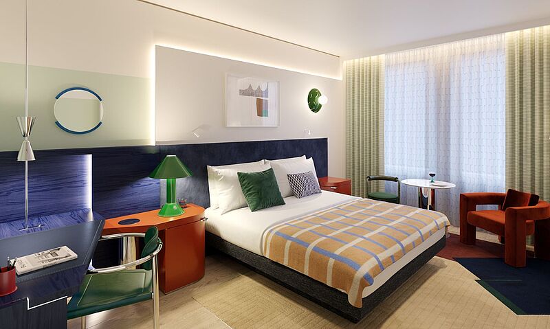 Blick in ein geräumiges Zimmer, links steht ein roter Nachttisch, ein blauer Schreibtisch und ein grüner Stuhl