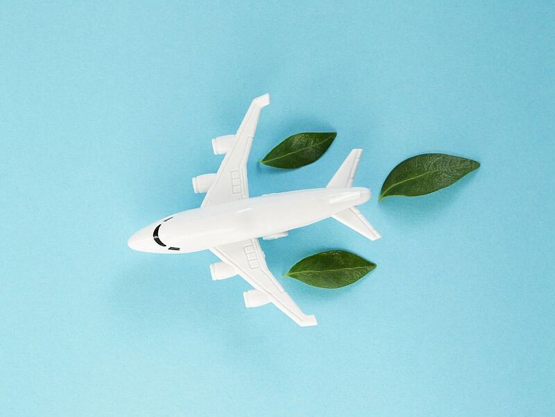 Ein weißes Spielzeugflugzeug auf hellblauem Hintergrund, umgeben von grünen Blättern, die aus den Triebwerken austreten – symbolisiert umweltfreundliches oder nachhaltiges Fliegen.