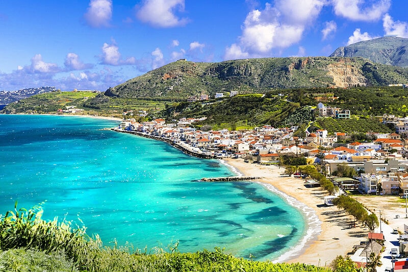 Küstenansicht von Kreta: Türkisblaues Meer, geschwungener Sandstrand, weiße Häuser mit roten Dächern vor grünen Berghängen