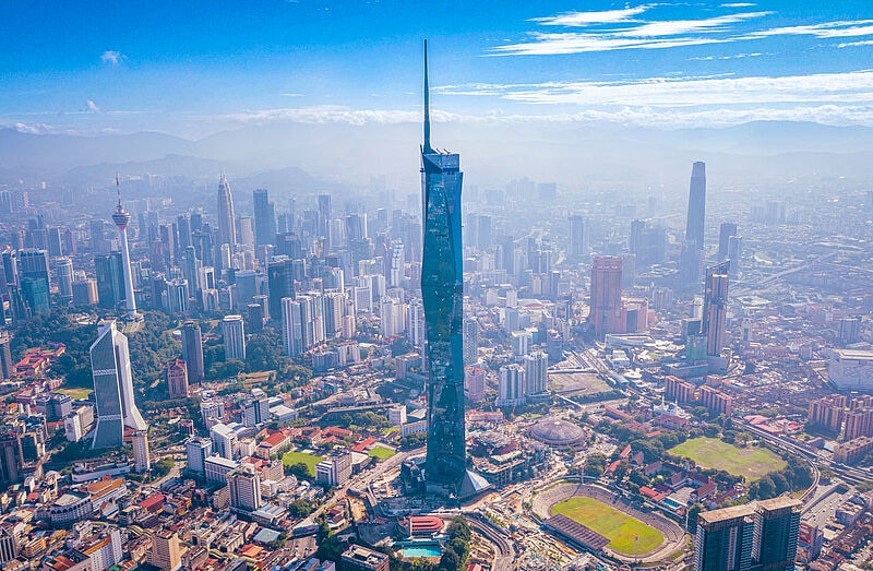Blick auf die Skyline von Kuala Lumpur, Malaysia, mit dem dominierenden Merdeka 118 Wolkenkratzer im Zentrum, umgeben von zahlreichen Hochhäusern, sportlichen Anlagen und grünen Flächen bei klarem, blauem Himmel.