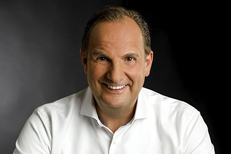 Ein Mann mit braunem, leicht zurückgekämmtem Haar trägt ein weißes Hemd mit offenem Kragen und sitzt vor einem dunklen, neutralen Hintergrund. Die Belichtung betont die linke Bildseite, während der Gesamteindruck seriös und professionell wirkt.