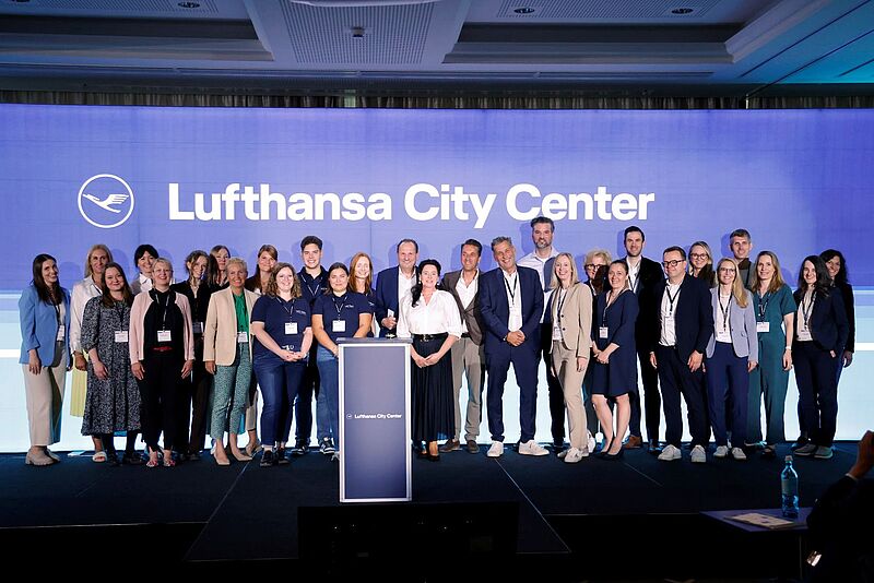 Eine große Gruppe von etwa 30 Personen steht auf einer Bühne vor einer blauen Wand mit dem Logo und Schriftzug „Lufthansa City Center“. Die Personen sind überwiegend geschäftlich gekleidet, einige tragen Firmenkleidung mit Namensschildern und Lanyards. In der Mitte steht ein Rednerpult mit dem Lufthansa City Center Logo. Die Stimmung wirkt professionell und zugleich kollegial. Die Veranstaltung findet in einem modernen Konferenzraum statt.