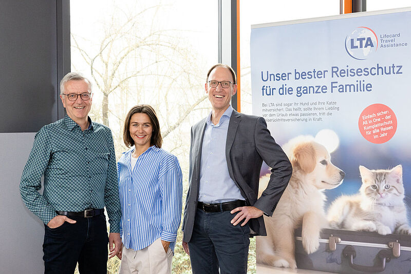 Drei Manager des Reiseversicherers LTA vor einem Werbebanner für Familienreiseschutz mit Haustierversicherung. Abgebildet sind dort ein Hund und eine Katze auf einem Reisekoffer.