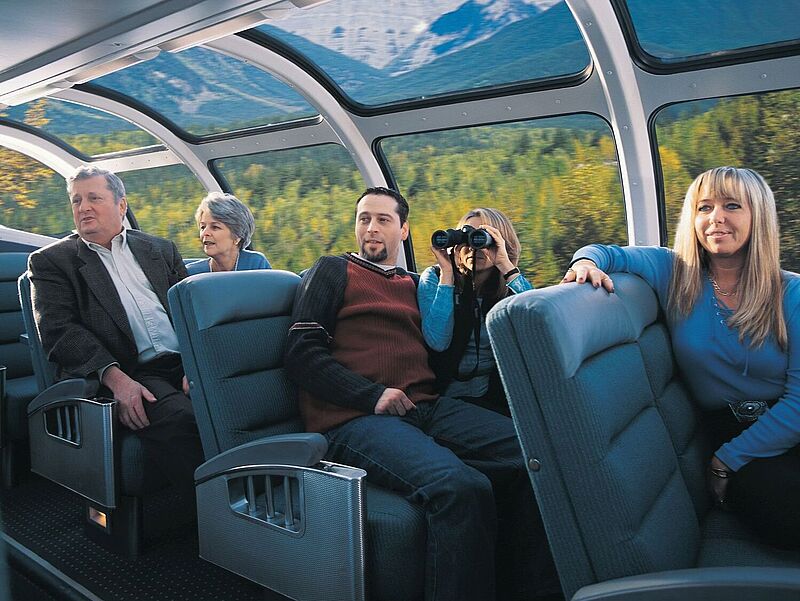 Eine Gruppe von Menschen sitzt entspannt in einem Panoramawagen eines Zuges mit großen, gewölbten Glasfenstern, durch die man eine malerische Landschaft mit Wäldern und Bergen sehen kann. Eine Frau benutzt ein Fernglas, um die Aussicht zu betrachten. Die Atmosphäre wirkt ruhig und genussvoll, während der Zug durch die Natur fährt.