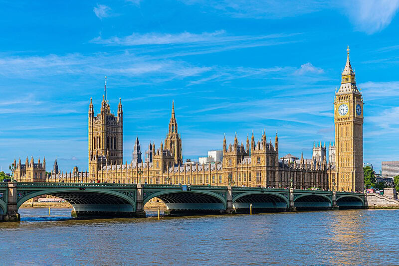 Der Westminster-Palast, der Big Ben und die Westminster-Brücke mit der Themse in London an einem sonnigen Morgen. Im Hintergrund ist blauer Himmel mit Schönwetterwolken zu sehen.