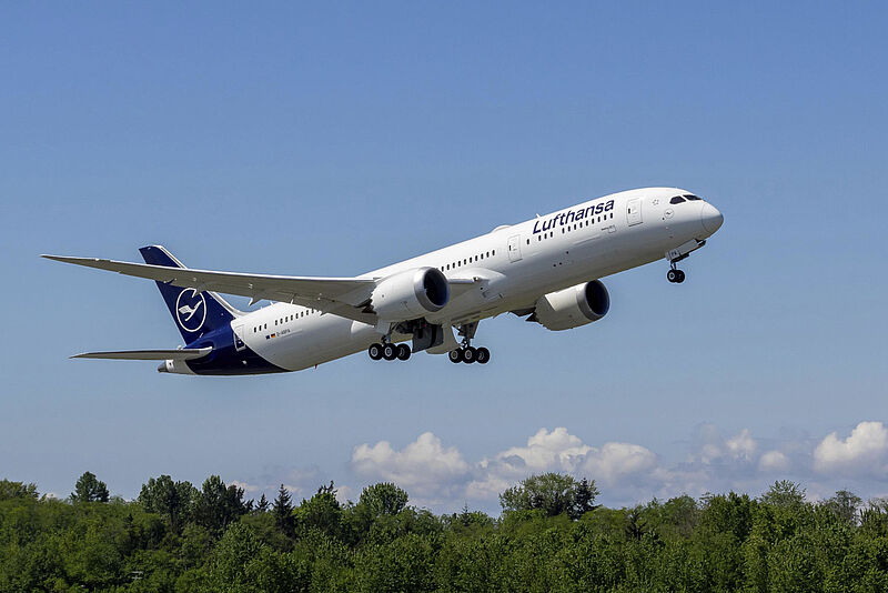 Ein Langstreckenjet des Typs Boeing 787 von Lufthansa hebt vor einem blauen Himmel und Bäumen ab.