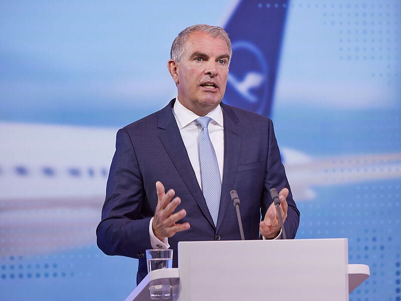 Ein Konzern-Manager mittleren Alters in dunklem Anzug, weißem Hemd und blauer Krawatte steht redend und gestikulierend hinter einem Rednerpult. Im Hintergrund ist unscharf auf einer Leinwand eine Heckflosse eines Lufthansa-Flugzeugs zu sehen.