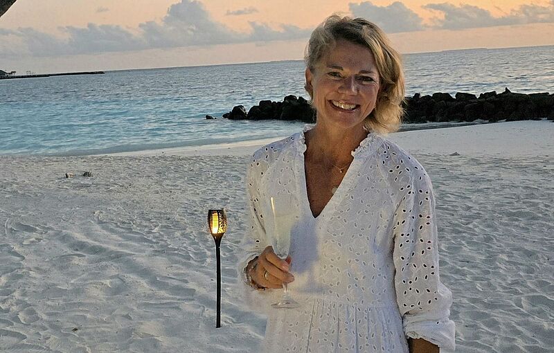 Eine lächelnde Frau steht barfuß am weißen Sandstrand bei Sonnenuntergang. Sie trägt ein luftiges, weißes Kleid mit Lochstickerei und hält ein Glas in der rechten Hand. Im Hintergrund leuchtet eine Fackel im Sand, das ruhige Meer glitzert in Blautönen, und dunkle Felsen ragen am Horizont ins Wasser. Der Himmel ist mit weichen Wolken in Pastellfarben gefärbt.