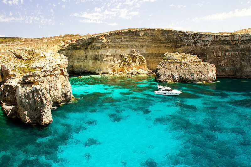 Türkisblaue Bucht umgeben von hohen Kalksteinklippen in Malta, ein weißes Motorboot ankert im kristallklaren Wasser.