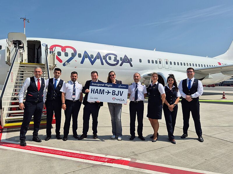 Eine Gruppe von neun Personen, bestehend aus Flugbegleitern, Piloten und Flughafenpersonal, steht vor einem Flugzeug der Fluggesellschaft MGA auf dem Rollfeld. Zwei Personen in der Mitte halten ein Schild mit der Aufschrift „Inaugural flight HAM → BJV, Juni 2025“. Im Hintergrund ist das Flugzeug mit MGA-Logo und türkischer Flagge am Leitwerk zu sehen. Der Anlass ist der Erstflug zwischen Hamburg (HAM) und Bodrum (BJV)