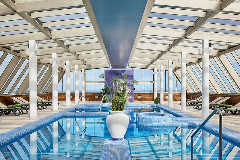 Heller, moderner Spa-Bereich mit überdachtem Innenpool im Hotel Melia Costa Atlantis auf Teneriffa. Der Raum ist von großen Glasfenstern umgeben, die Tageslicht einlassen, und mit weißen Säulen, Liegestühlen und einem zentralen Pflanzgefäß im Pool stilvoll gestaltet