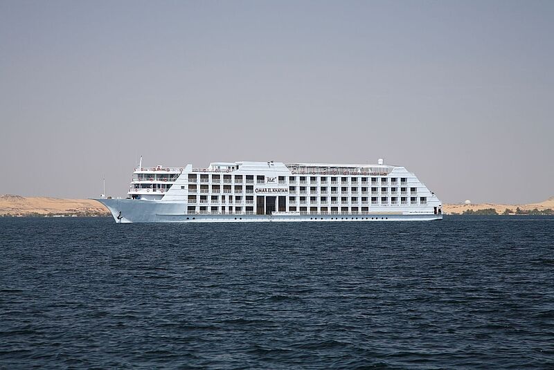 Ein großes, weißes Flusskreuzfahrtschiff mit dem Namen „Omar El Khayam“ fährt auf dem Nil. Das Schiff hat mehrere Decks mit Balkonen und Rettungsringen an der Reling. Im Hintergrund sind sandige Dünen und die karge Landschaft Oberägyptens unter einem klaren Himmel zu sehen.
