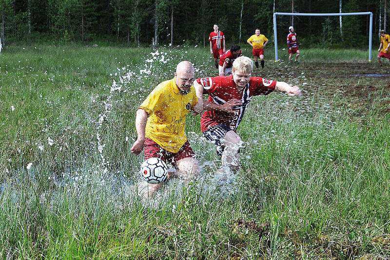 Männer in roten und gelben Trikots spielen auf einer matschigen Wiese mit hohem Gras und Pfützen Fußball