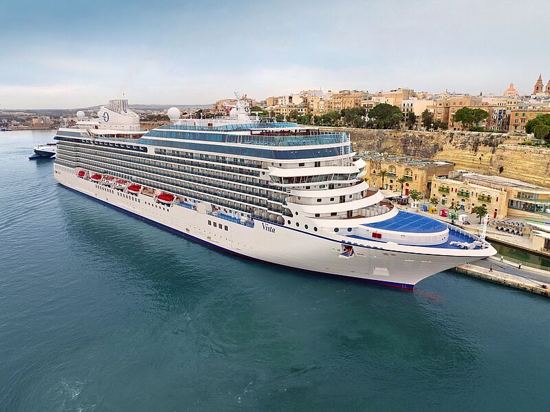 Ein hochaufragendes Kreuzfahrtschiff liegt an einem Kai, dahinter die nicht so hohen Gebäude einer Stadt.