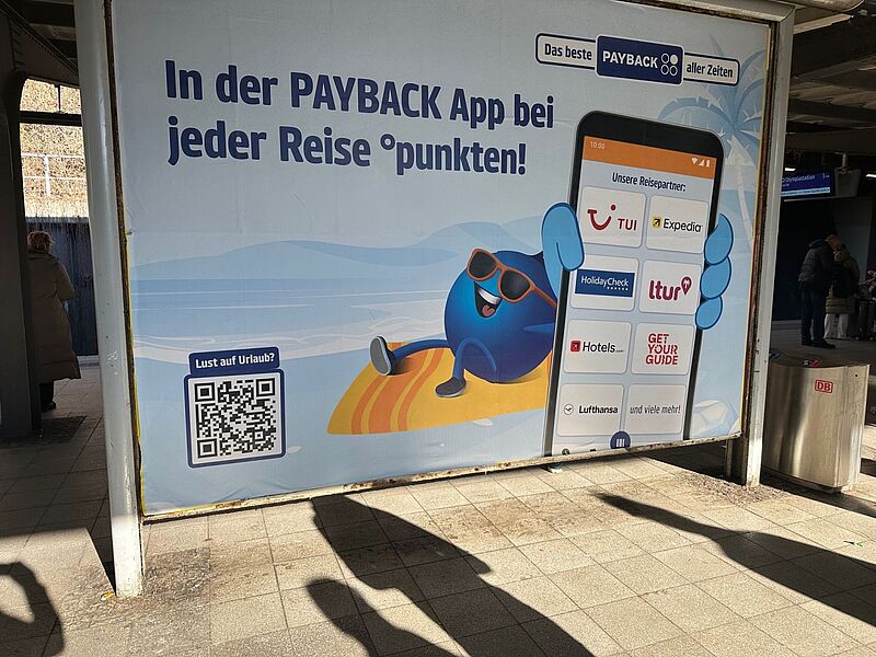 Werbeplakat an einem Bahnhof für die PAYBACK App mit dem Slogan „In der PAYBACK App bei jeder Reise punkten!“. Das Plakat zeigt ein Cartoon-Maskottchen mit Sonnenbrille auf einem Liegestuhl am Strand. Daneben ist ein großes Smartphone abgebildet, das Reiseanbieter wie TUI, Expedia, HolidayCheck, ltur, Hotels.com, GetYourGuide und Lufthansa zeigt. Unten links befindet sich ein QR-Code mit dem Text „Lust auf Urlaub?“. Rechts im Bild sind Fahrgäste und das DB-Logo zu sehen.