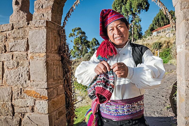 Ein freundlicher Mann mit andinischen Gesichtszügen strickt mit einem Nadelspiel. Er hat eine gestrickte Mütze auf. Er wohnt auf der Insel Taquile, die im Titicacasee liegt. Dort ist Stricken Männersache. Die Insel ist gebirgig und die Männer stricken auch, wenn sie die Pfade hoch und runter gehen. 