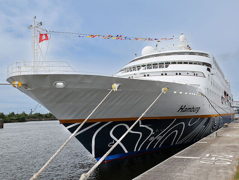 An einer Pier liegt ein Kreuzfahrtschiff mit dunkelblauem Bug und weißem Aufbau. Am Bug prangt in weißer Schrift der Schiffsname Hamburg.