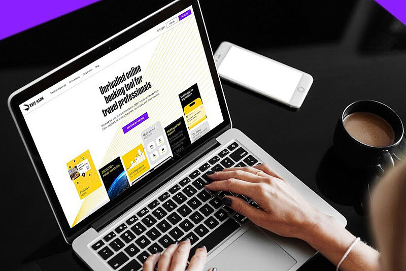 Person arbeitet an einem Laptop, auf dem eine Website für ein Online-Buchungstool für Reiseprofis angezeigt wird; daneben liegen ein Smartphone und eine Tasse Kaffee auf einem schwarzen Tisch