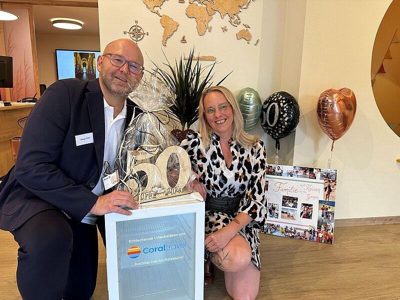 Ein Mann im Anzug und eine Frau im gemusterten Kleid knien lächelnd nebeneinander in einem Büro. Sie halten eine dekorative Zahl ‚50‘ mit der Jahreszahl 1975–2025. Im Hintergrund sind Ballons mit der Aufschrift ‚50‘, eine Fotocollage und eine Weltkarte an der Wand zu sehen.