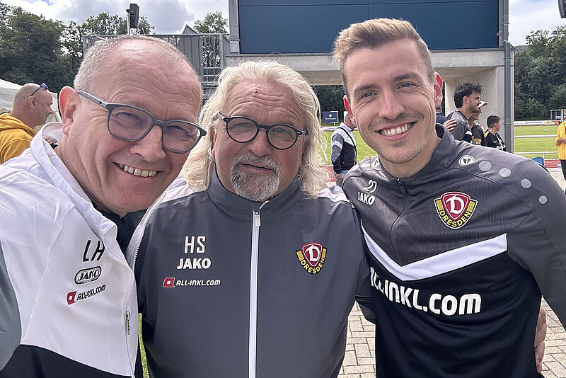 Drei Personen in JAKO-Trainingskleidung mit Dynamo Dresden Logo und ALL-INKL.com Sponsoring vor einer Anzeigetafel im Stadion.