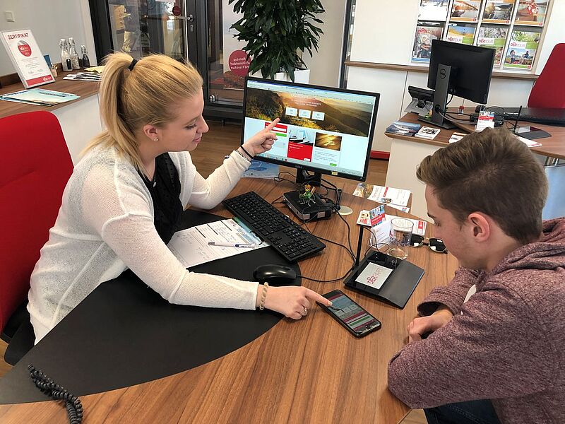 Reisebüro-Mitarbeiterin berät jungen Kunden am Schreibtisch mit Computer und Smartphone – persönliche Urlaubsberatung im modernen Reisebüro