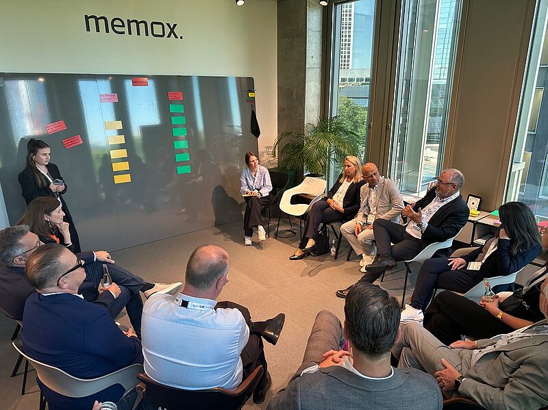 Eine Gruppe von etwa 15 Personen sitzt in einem modernen, lichtdurchfluteten Besprechungsraum im Kreis und diskutiert. An der Wand hängt eine große magnetische Tafel mit farbigen Notizzetteln. Zwei Frauen stehen bzw. sitzen an der Tafel, während ein Mann spricht und die Aufmerksamkeit der Gruppe auf sich zieht. Im Hintergrund sind große Fenster mit Blick auf eine Stadtlandschaft zu sehen. Das Logo