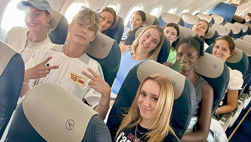 Eine Gruppe junger Menschen sitzt lächelnd in den Sitzen eines Flugzeugs. Sie wirken fröhlich und entspannt, einige zeigen das Peace-Zeichen. Die Kabine ist hell und freundlich beleuchtet, im Hintergrund sind weitere Fluggäste zu sehen.