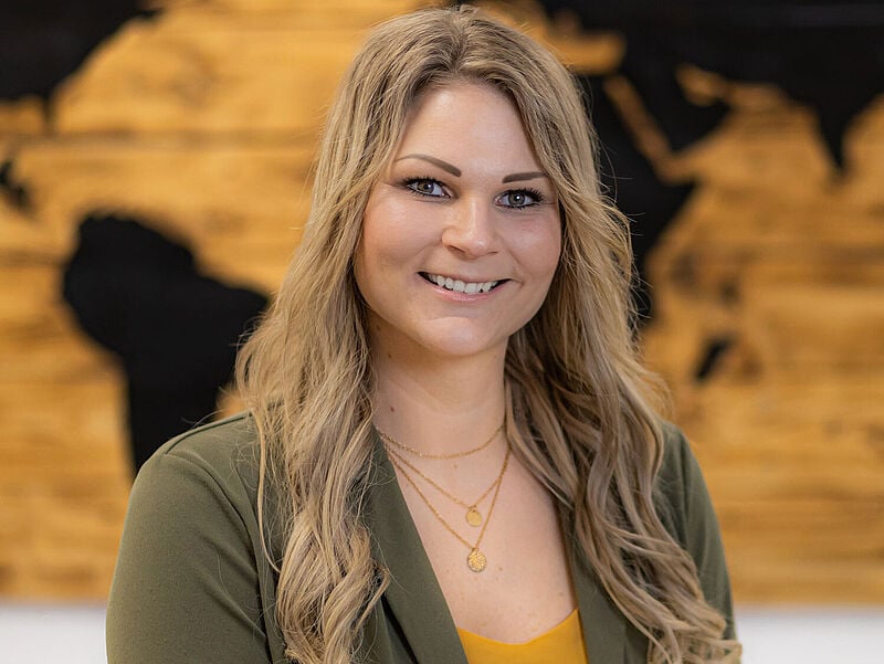 Junge Reisebüro-Inhaberin mit langen dunkelblonden Haaren lächelt in die Kamera, im Hintergrund hängt eine Weltkarte