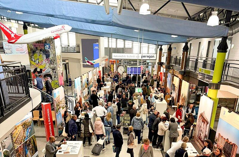 Blick auf eine gut besuchte Reisemesse mit zahlreichen Informationsständen, bunten Reiseplakaten und Modellen von Flugzeugen über den Ständen. Besucher schlendern durch die Gänge, führen Gespräche mit Ausstellern und informieren sich über Reiseangebote. Die Atmosphäre wirkt lebendig und interessiert.