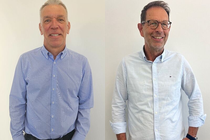 Zwei Männer stehen nebeneinander vor einem hellen Hintergrund. Der Mann links trägt ein hellblau kariertes Hemd mit Knopfleiste und hat kurzes graues Haar. Der Mann rechts trägt ein hellblaues Hemd mit aufgerollten Ärmeln und eine Brille, er hat ebenfalls graue Haare und lächelt in die Kamera.