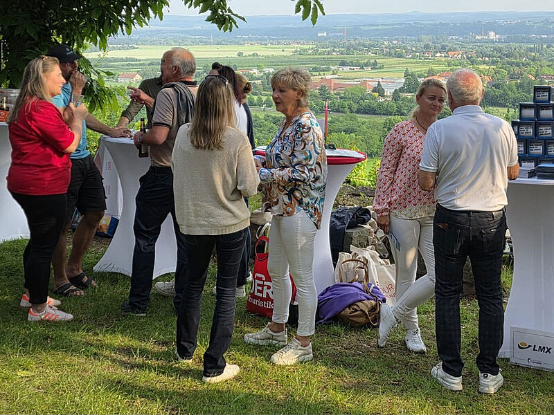 Eine Gruppe von Menschen steht im Freien an Stehtischen und unterhält sich bei einem Treffen oder Event. Im Hintergrund ist eine weite Landschaft mit Feldern, Bäumen und Gebäuden zu sehen. Auf einem der Tische sind gestapelte kleine Kartons und Prospekte mit einem Schild des Reiseveranstalters LMX. Einige Taschen am Boden tragen Logos von DERTOUR GROUP und Eberhardt Travel, was auf eine Veranstaltung mit touristischen Partnern hinweist.