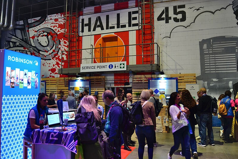 Industrielle Messehalle mit Service Point, Robinson-Messestand mit blauem Display und wartenden Besuchern vor verschiedenen Ausstellerständen