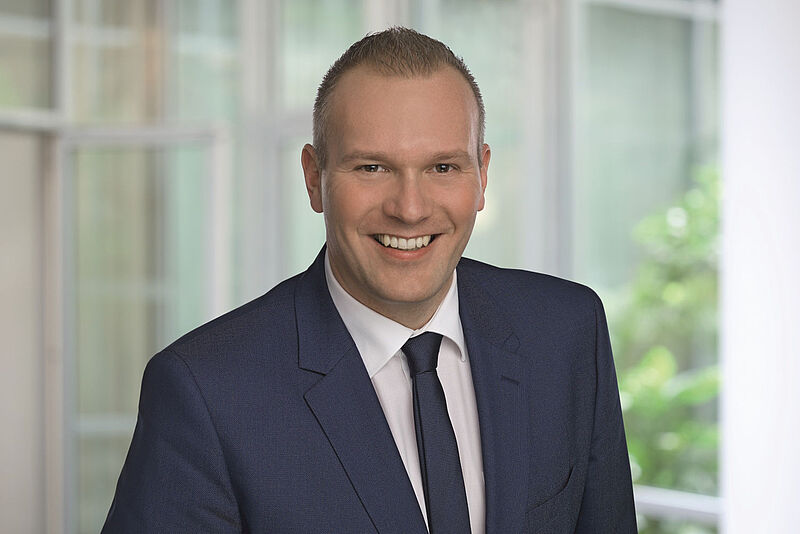 Professionelles Foto eines Managers mit grauem Anzug und weißen Hemd, der in die Kamera lächelt. Die Person ist durch einen unscharfen hellen Hintergrund mit einem Fenster und einem Baum freigestellt.