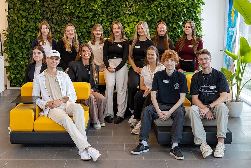 Vierzehn junge Auszubildende posieren in zwei Reihen in einem modernen Bürobereich vor einer grünen Pflanzenwand; einige sitzen auf gelben und grauen Sesseln, andere stehen dahinter, alle lächeln in die Kamera.