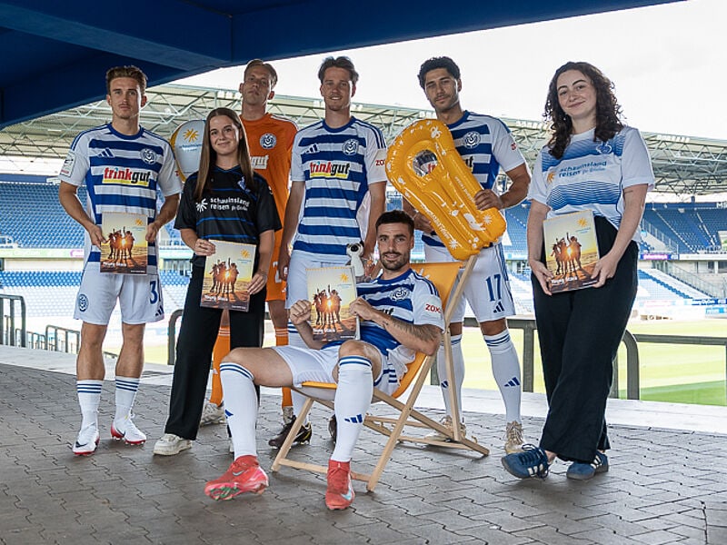 Eine Gruppe von sechs Personen posiert im Stadion des MSV Duisburg. Vier Fußballspieler in weiß-blau gestreiften Trikots und ein Torwart im orangefarbenen Trikot stehen zusammen mit zwei Frauen, die Fan-Shirts tragen. Ein Spieler sitzt entspannt in einem Liegestuhl. Mehrere Personen halten Poster mit einem Sommermotiv, ein Spieler hält zusätzlich eine große gelbe Luftmatratze in Form einer „9“. Im Hintergrund sind die leeren Tribünen des Stadions zu sehen.