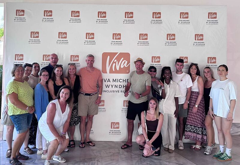 Eine Gruppe von etwa fünfzehn Personen steht vor einer Fotowand mit dem Logo 