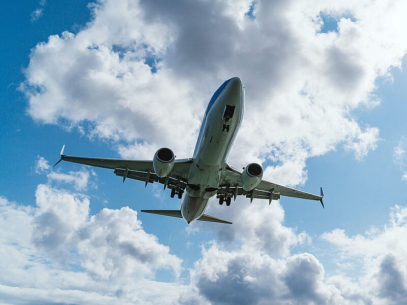 Ein Verkehrsflugzeug im Landeanflug vor einem blauen, bewölkten Himmel. Blick von unten auf den Flugzeugrumpf mit ausgefahrenem Fahrwerk und Landeklappen.