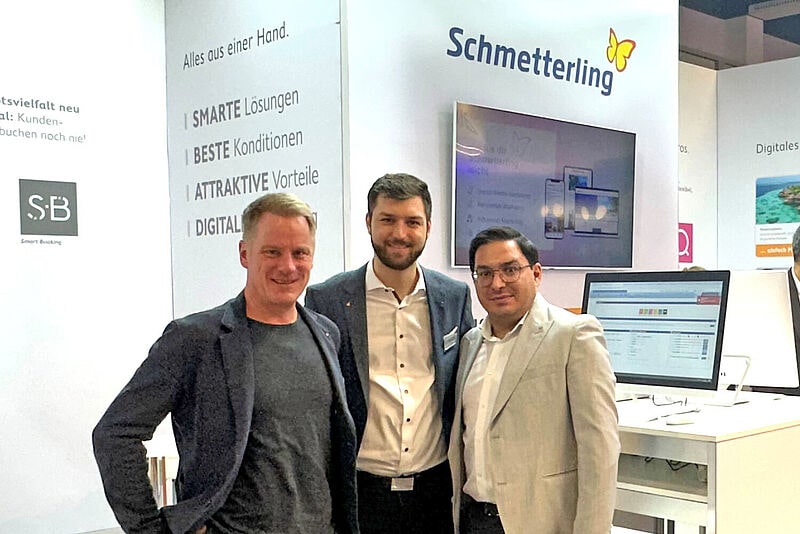 Drei Männer posieren lächelnd für ein Foto auf der Tourismusmesse ITB 2025 am Stand des Unternehmens Schmetterling. Im Hintergrund sind Monitore, Informationswände und weitere Messebesucher zu sehen. Der Messestand ist modern gestaltet mit dem Logo und Slogan des Unternehmens.