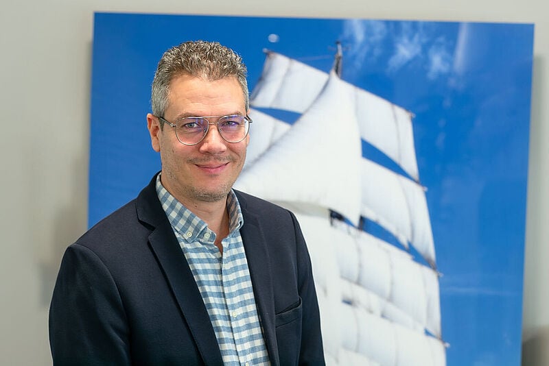 Ein Vertriebler Ende 40 mit grauem, gelocktem Haar und Brille lächelt freundlich in die Kamera. Er trägt ein blauweiß kariertes Oberhemd und ein dunkles Sakko, im Hintergrund ist ein mannshohes Bild von einem Segelschiff in Teilen zu erkennen.