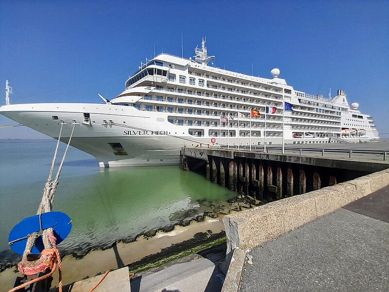 Das Kreuzfahrtschiff Silver Moon mit hellgrau-weißem Rumpf liegt vertäut an einer beflaggten Pier. 