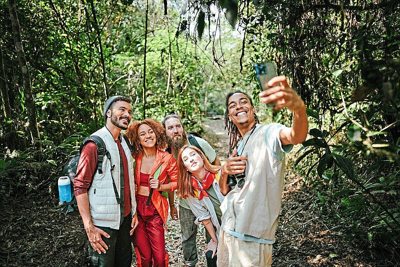 Gruppe von fünf Wanderern verschiedener Ethnien macht ein fröhliches Selfie auf einem Waldweg, ausgestattet mit Wanderausrüstung und Rucksäcken
