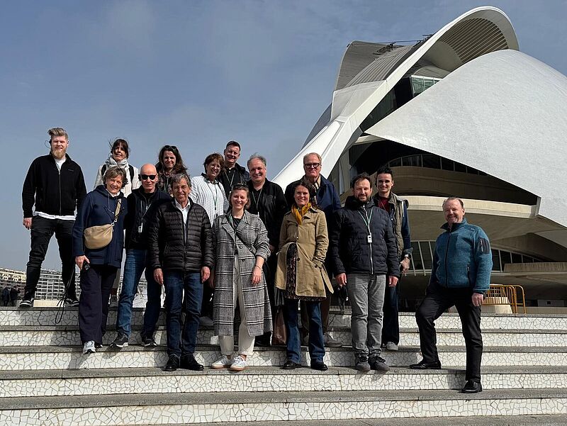 Eine Gruppe von 15 Personen steht auf einer Treppe vor einem modernen, weißen Gebäude mit geschwungener Architektur. Die Personen tragen Freizeitkleidung, einige mit Jacken und Schals. Sie lächeln in die Kamera, während sie sich für ein Gruppenfoto positionieren. Der Himmel ist blau mit wenigen Wolken, und im Hintergrund sind weitere Gebäude zu sehen.