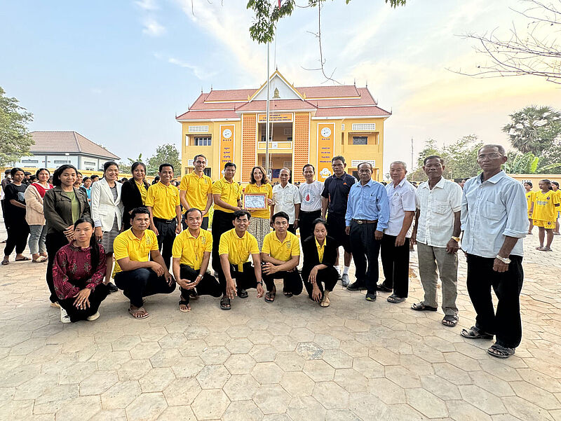 Eine große Gruppe von Menschen, bestehend aus Männern und Frauen unterschiedlichen Alters, steht auf einem Schulhof vor einem gelben Schulgebäude mit rotem Dach. Einige Personen tragen gelbe Shirts, andere sind formell gekleidet. In der Mitte der Gruppe wird eine Urkunde präsentiert. Im Hintergrund sind weitere Personen sowie Kinder in gelber Schuluniform zu sehen. Der Himmel ist leicht bewölkt und es herrscht eine freundliche Atmosphäre.