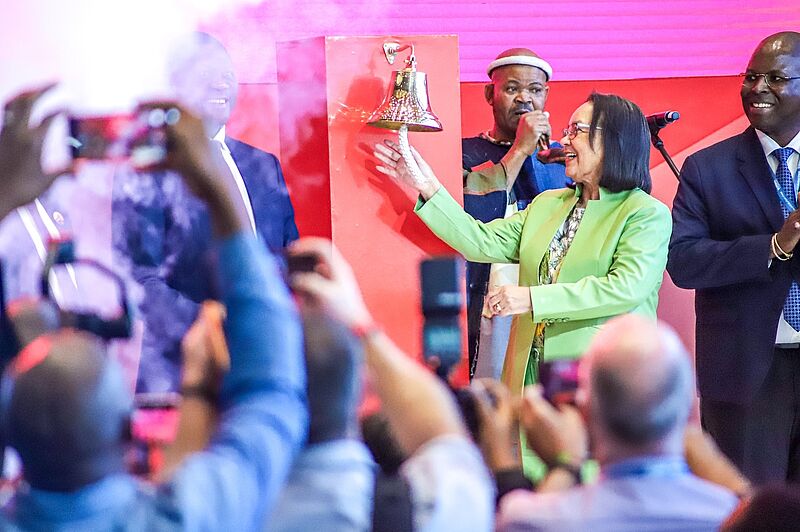 Person in grünem Blazer läutet eine Glocke auf einer Bühne vor fotografierendem Publikum, begleitet von Würdenträgern