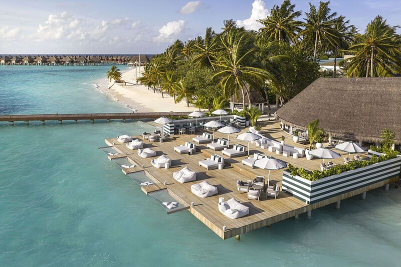 Luftaufnahme eines luxuriösen Malediven-Resorts mit Overwater-Bungalows, Holzterrasse mit weißen Liegen und Sonnenschirmen über türkisblauem Wasser und Palmen am Strand